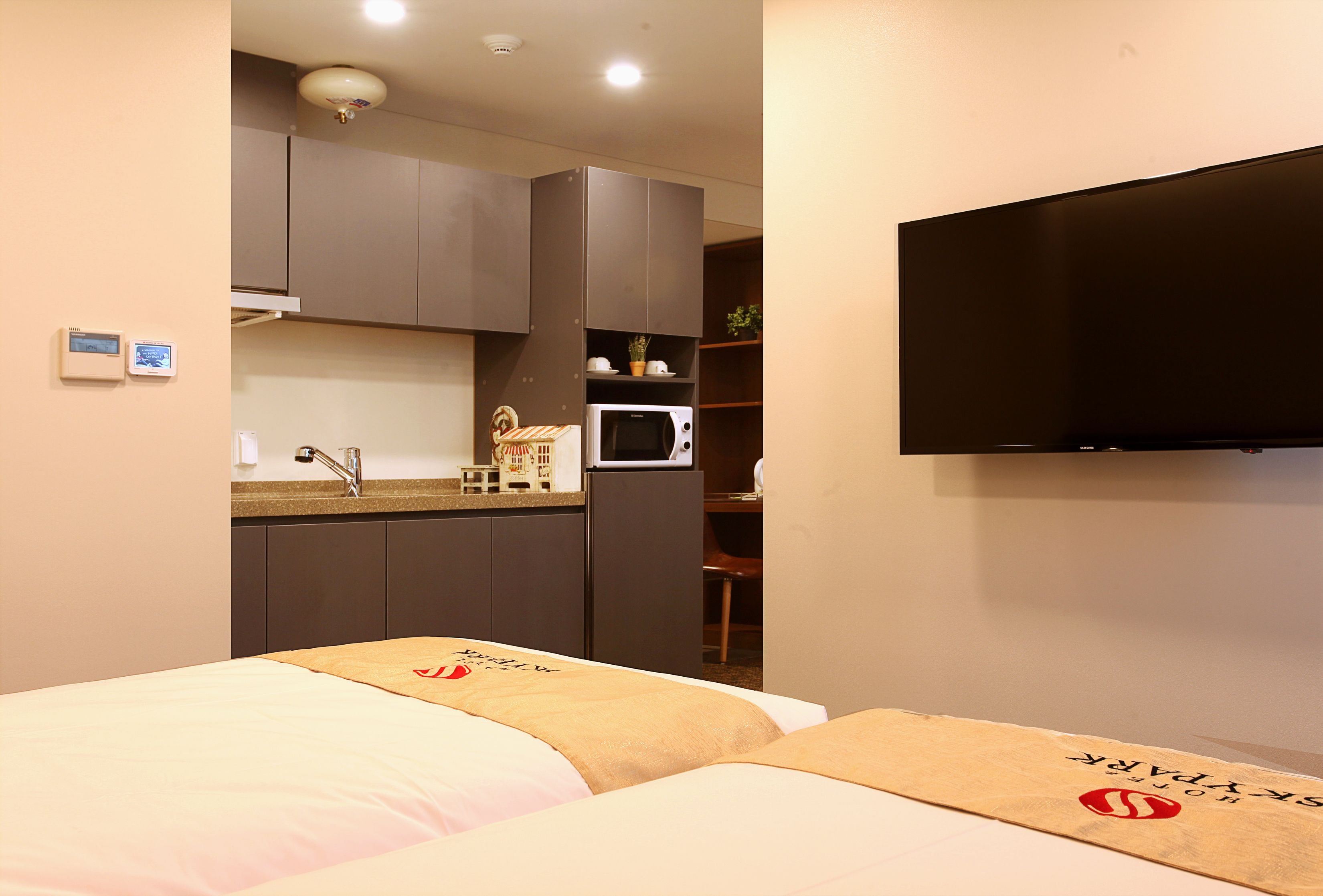 Quarto de hotel compacto com cozinha compacta, uma cama com cabeceira decorativa e uma televisão de tela plana fixada na parede.