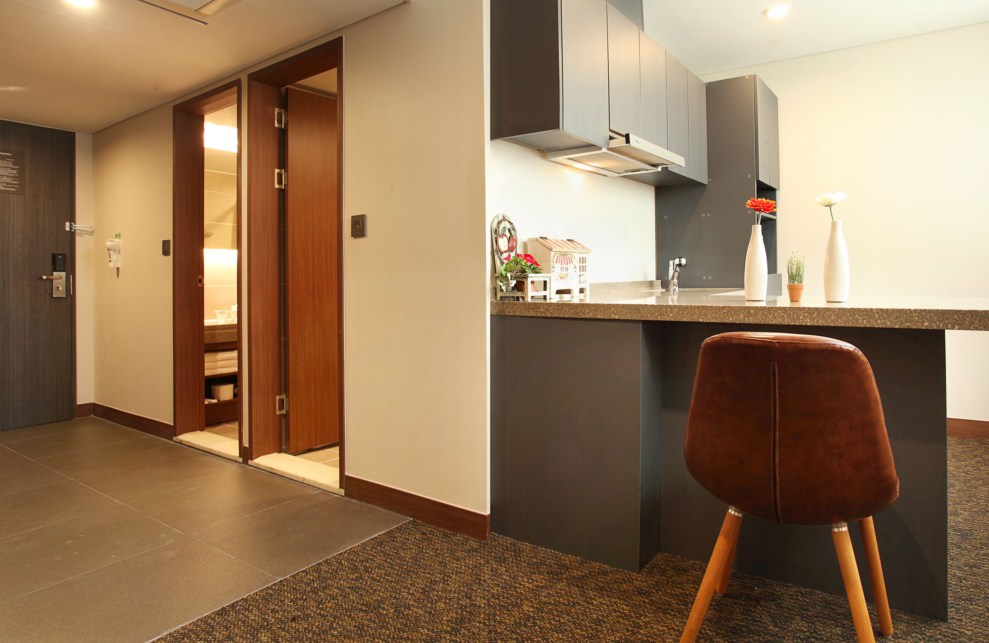 A modern hotel room with a wooden door leading to a bathroom, a kitchenette with a countertop, and a brown chair in the foreground.