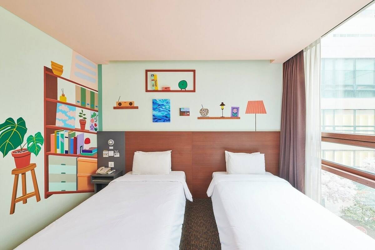 Hotellihuone, jossa on kaksi sänkyä, kirjahylly, työpöytä lampun kanssa, kello ja ikkuna, jossa on verhot.