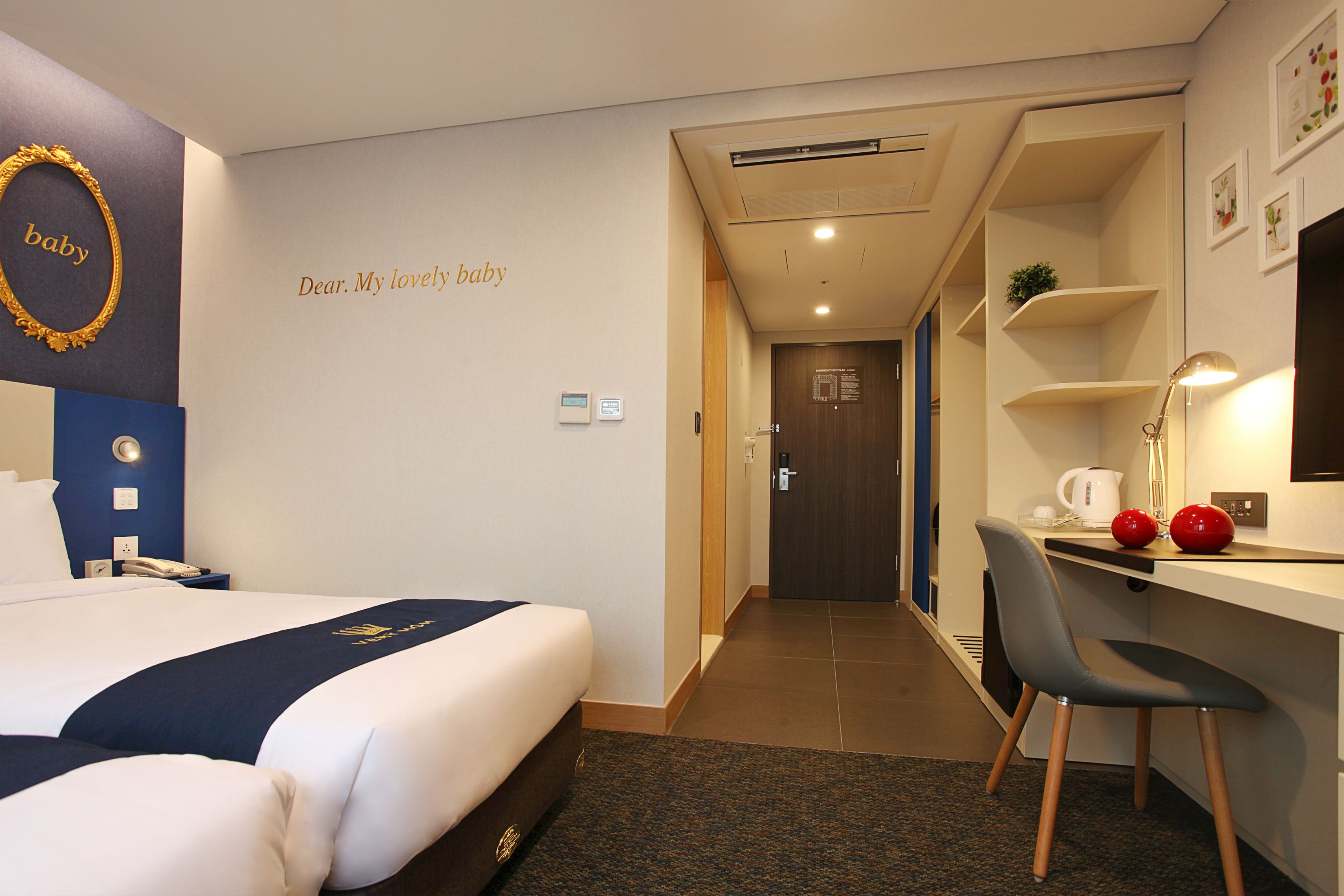 A hotel room with a bed, a desk, a chair, a desk lamp, and a wall with a decorative gold frame and a framed message.