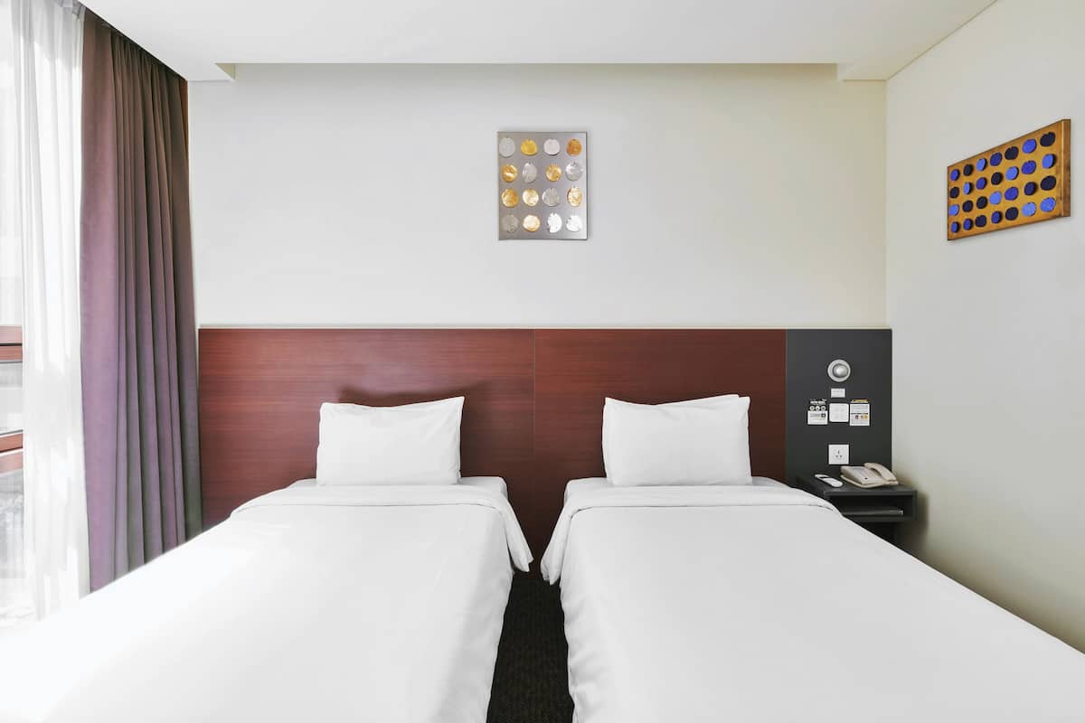 Hotellihuone, jossa on kaksi sänkyä, puinen pääty, seinään kiinnitetty puhelin ja ikkuna, jossa on verhot.