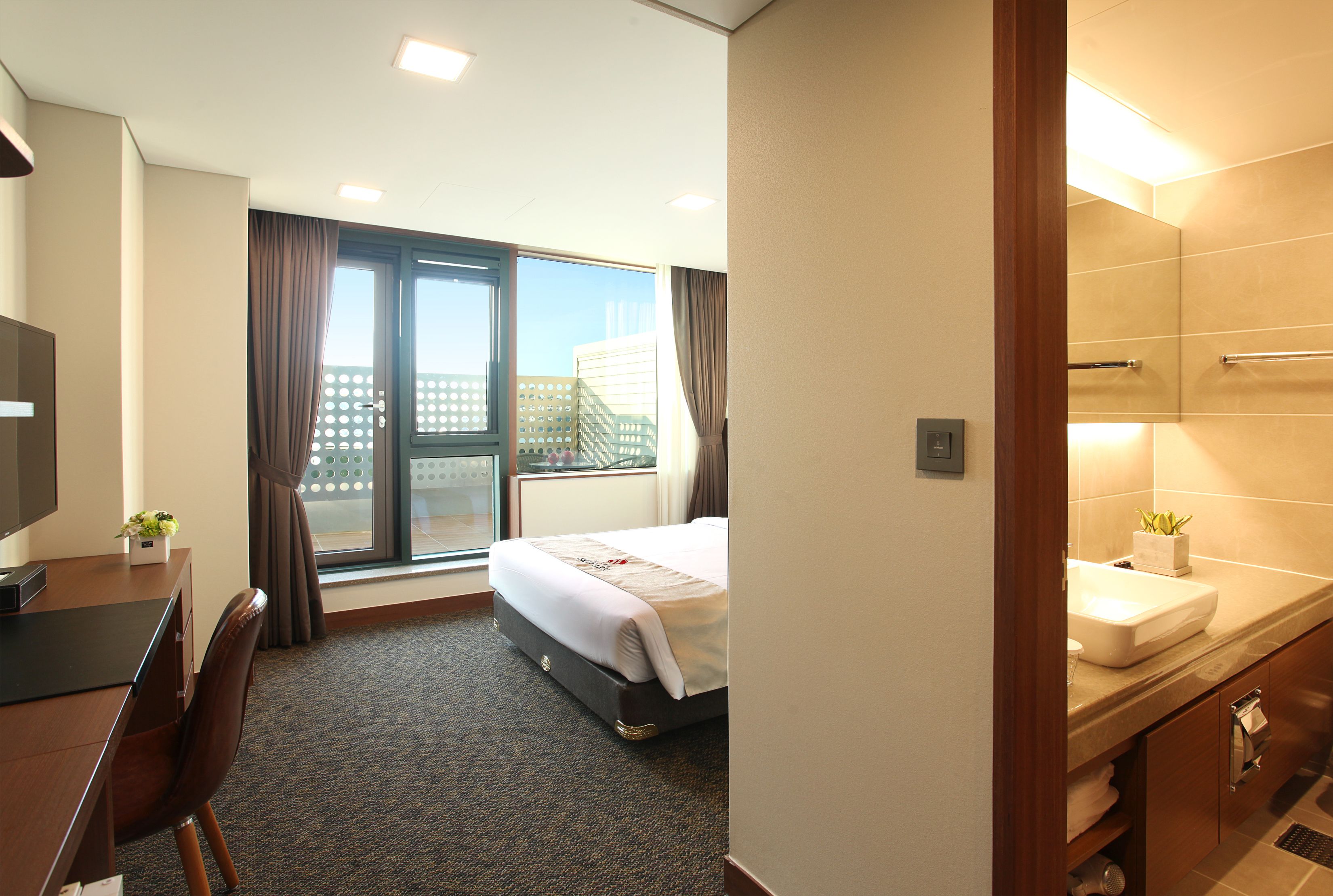 A hotel room with a bed, a desk, a chair, a TV, a window with curtains, and a bathroom with a sink and mirror.