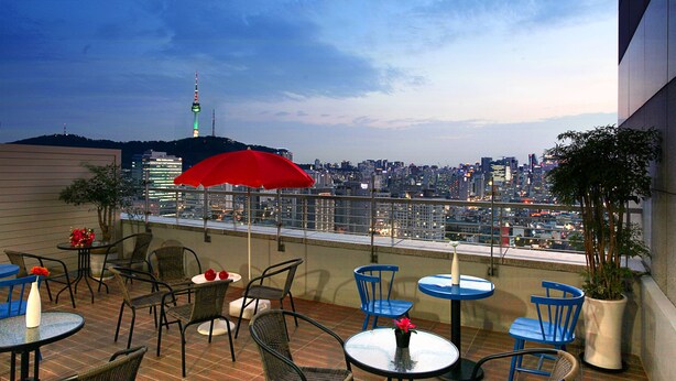 A rooftop terrace with tables and chairs, a red umbrella, and a cityscape view. - Hotel Skypark Kingstown Dongdaemun (Seoul)