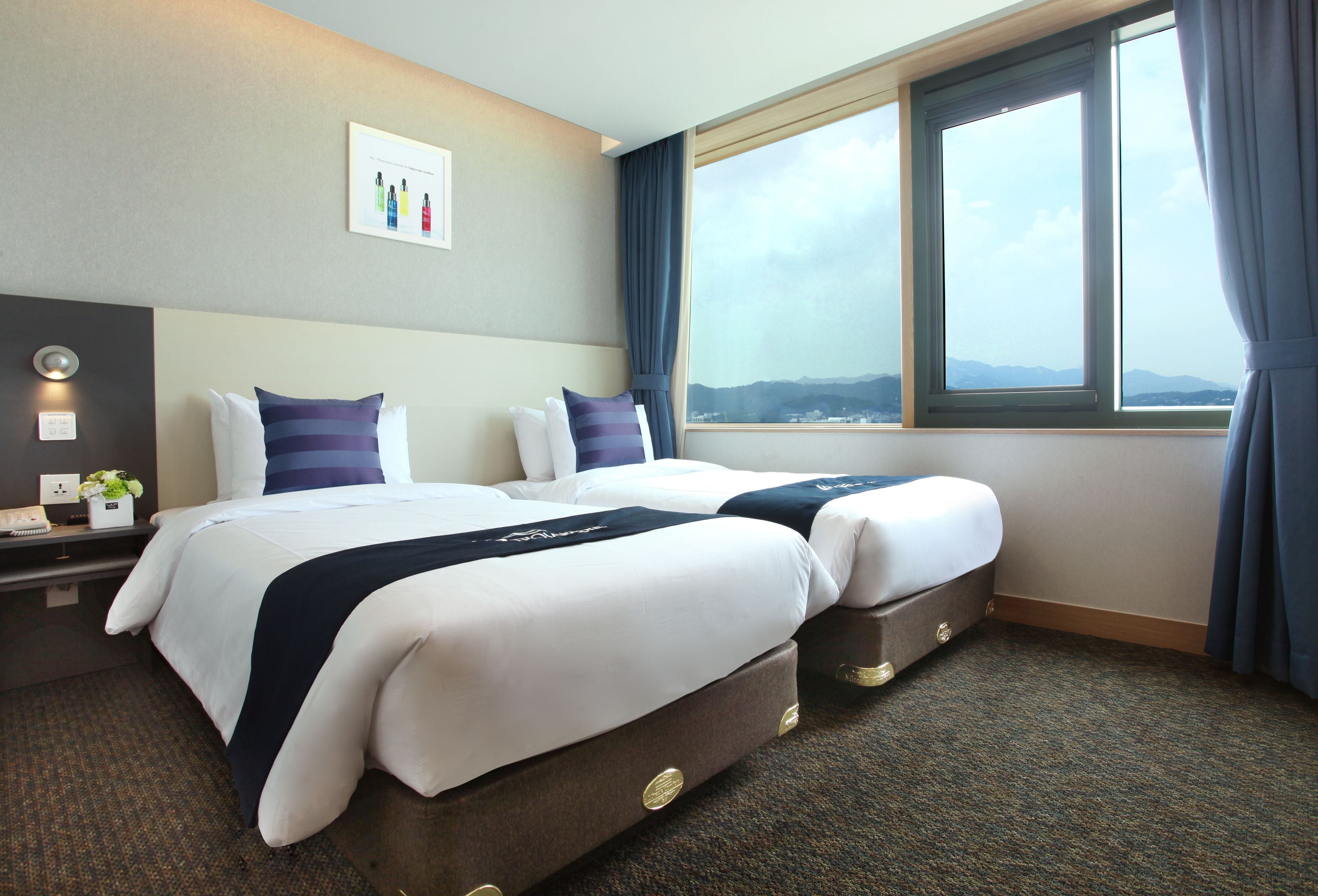 A hotel room with two beds, a large window with curtains, a bedside table with a lamp, and a wall-mounted artwork.