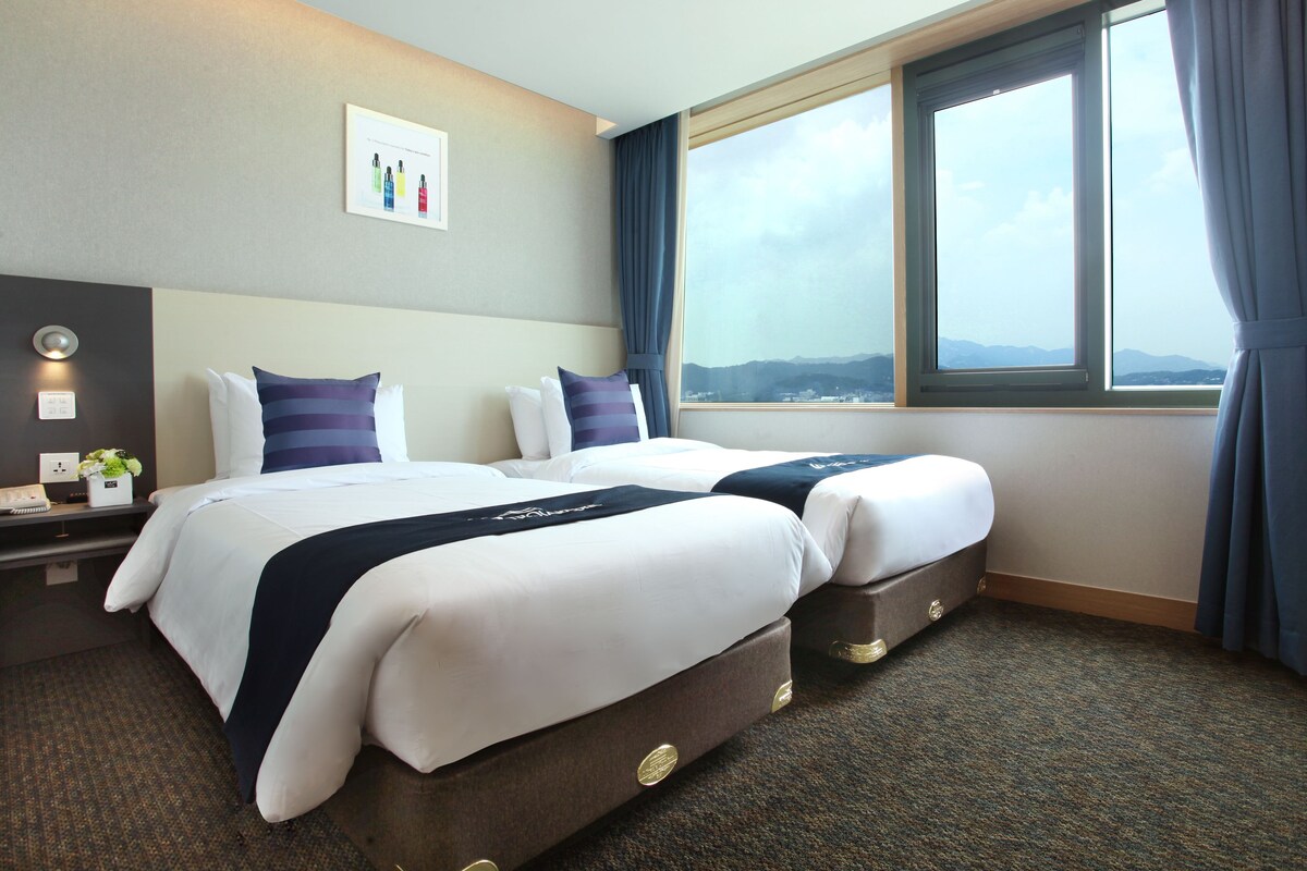 A hotel room with two beds, a large window with curtains, a bedside table with a lamp, and a wall-mounted artwork.