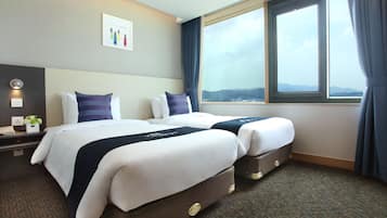 A hotel room with two beds, a large window with curtains, a bedside table with a lamp, and a wall-mounted artwork.