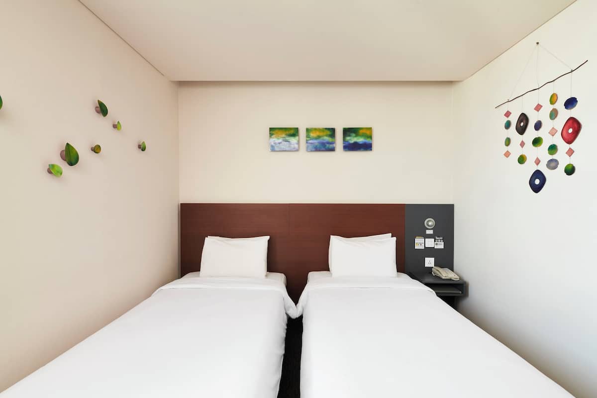 Hotellihuone, jossa on kaksi erillistä sänkyä, puinen pääty, pieni työpöytä, jossa on puhelin, sekä seinäkoristeita, kuten lehtiaiheisia koristeita ja kolme kehystettyä kuvaa.