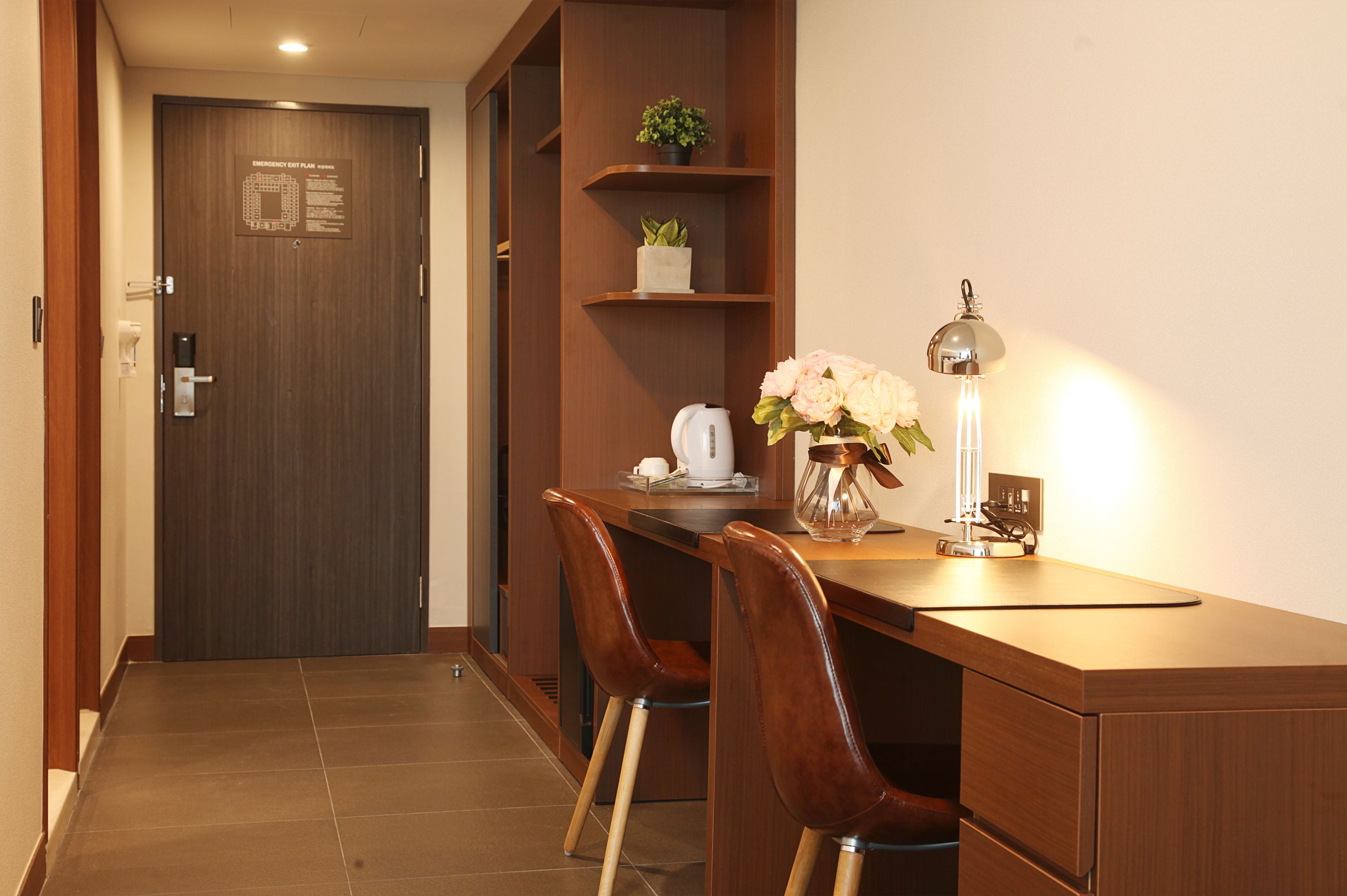 Quarto de hotel com uma escrivaninha de madeira, duas cadeiras, cafeteira, planta em vaso, um vaso com flores e uma luminária.