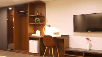 A modern hotel room with a wooden desk, a television, a small refrigerator, and a closet with shelves.