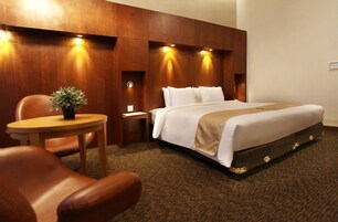 A hotel room with a large bed, a wooden nightstand, a small table with a plant, and leather chairs.