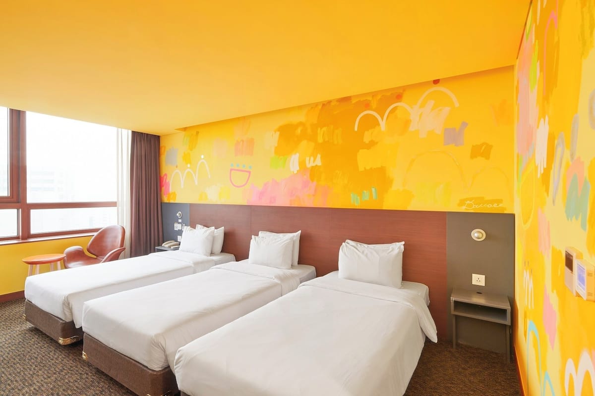 Hotellihuone, jossa on kolme erillistä sänkyä, pieni työpöytä, tuoli ja ikkuna, jossa on verhot.