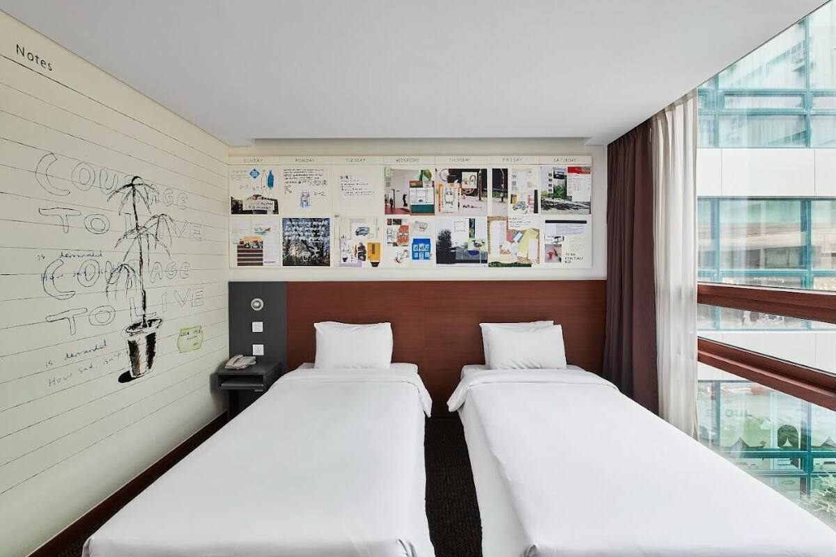 Hotellihuone, jossa on kaksi erillistä sänkyä, suuri ikkuna, josta on näkymä, ja seinä, joka on koristeltu erilaisilla muistiinpanoilla ja piirroksilla.