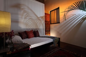 Ein Schlafzimmer mit einem Bett, Kissen, einem Nachttisch mit Lampe und Büchern, einer dekorativen Wandverkleidung und einer großen Pflanze.