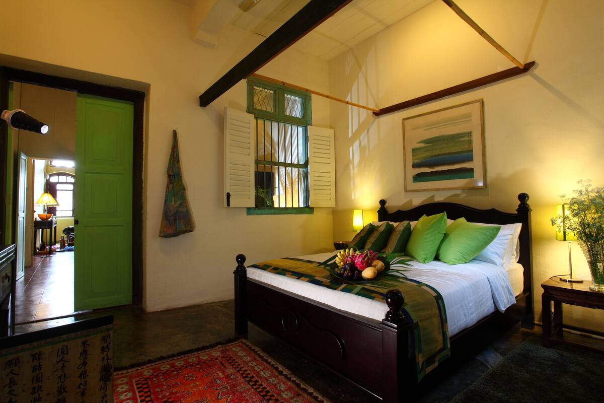 Um quarto com uma cama, uma porta verde, uma janela com persianas, um quadro na parede e um abajur em uma mesa de cabeceira.