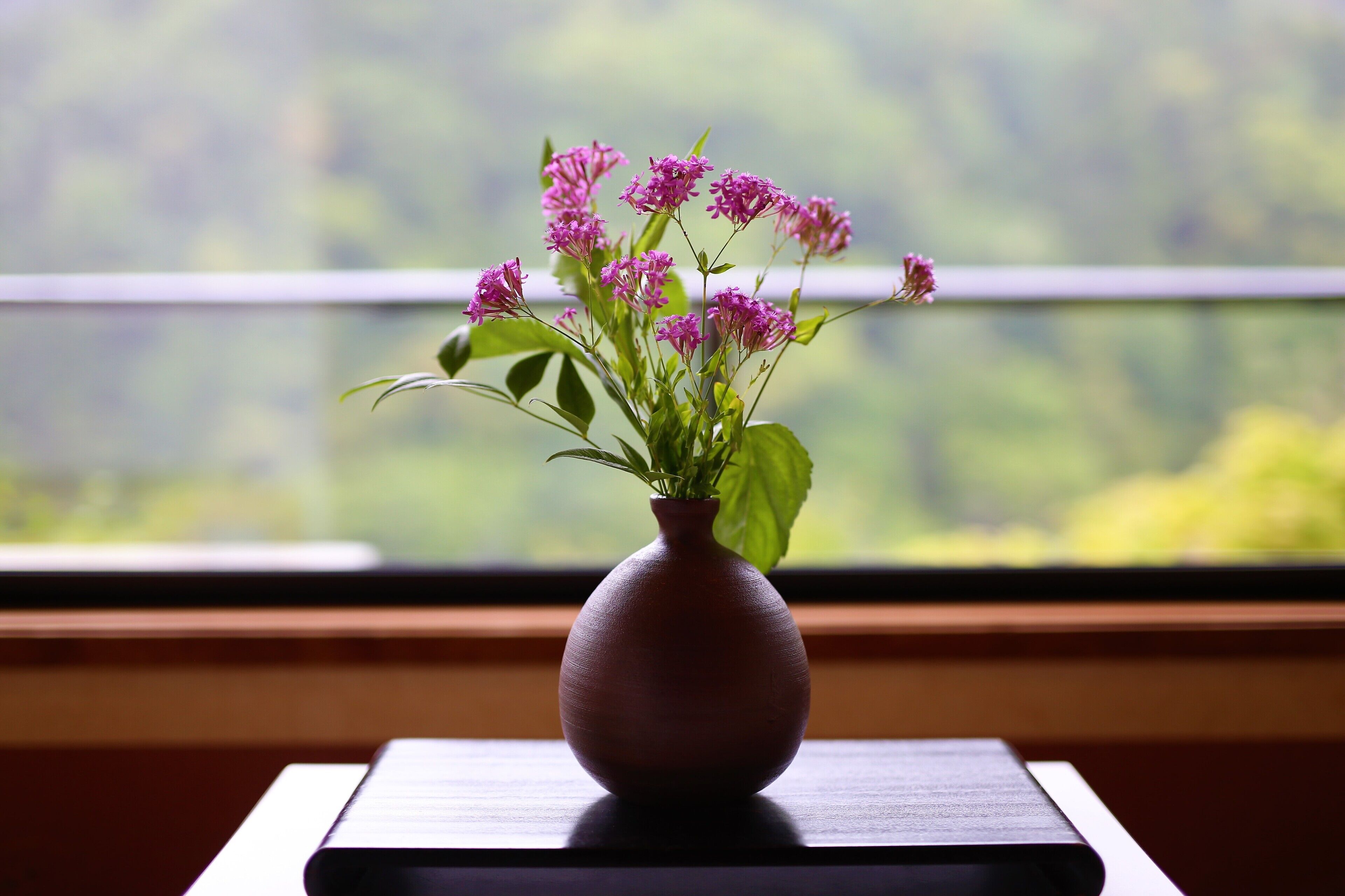 Eine Vase mit rosa Blumen auf einem Tisch neben einem Fenster.