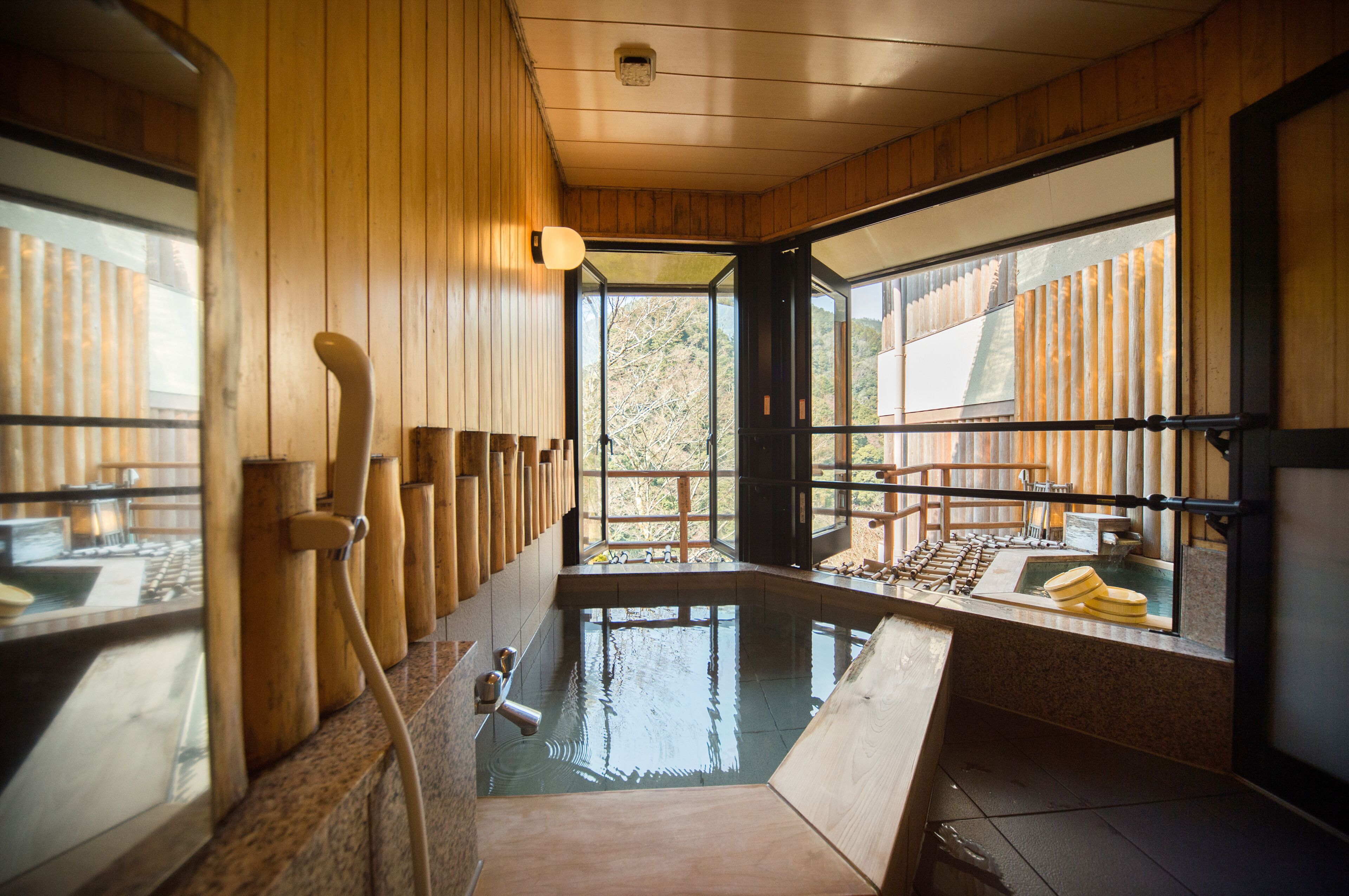 Uma onsen japonesa tradicional com interiores de madeira, um banho de águas termais e vista para a floresta circundante.
