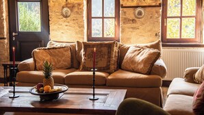 A cozy room with a stone wall, a wooden door, and a window with a view of trees. There's a sofa with cushions and a table with a fruit bowl and candles. - Epoches Luxury Suites (Karpenisi)