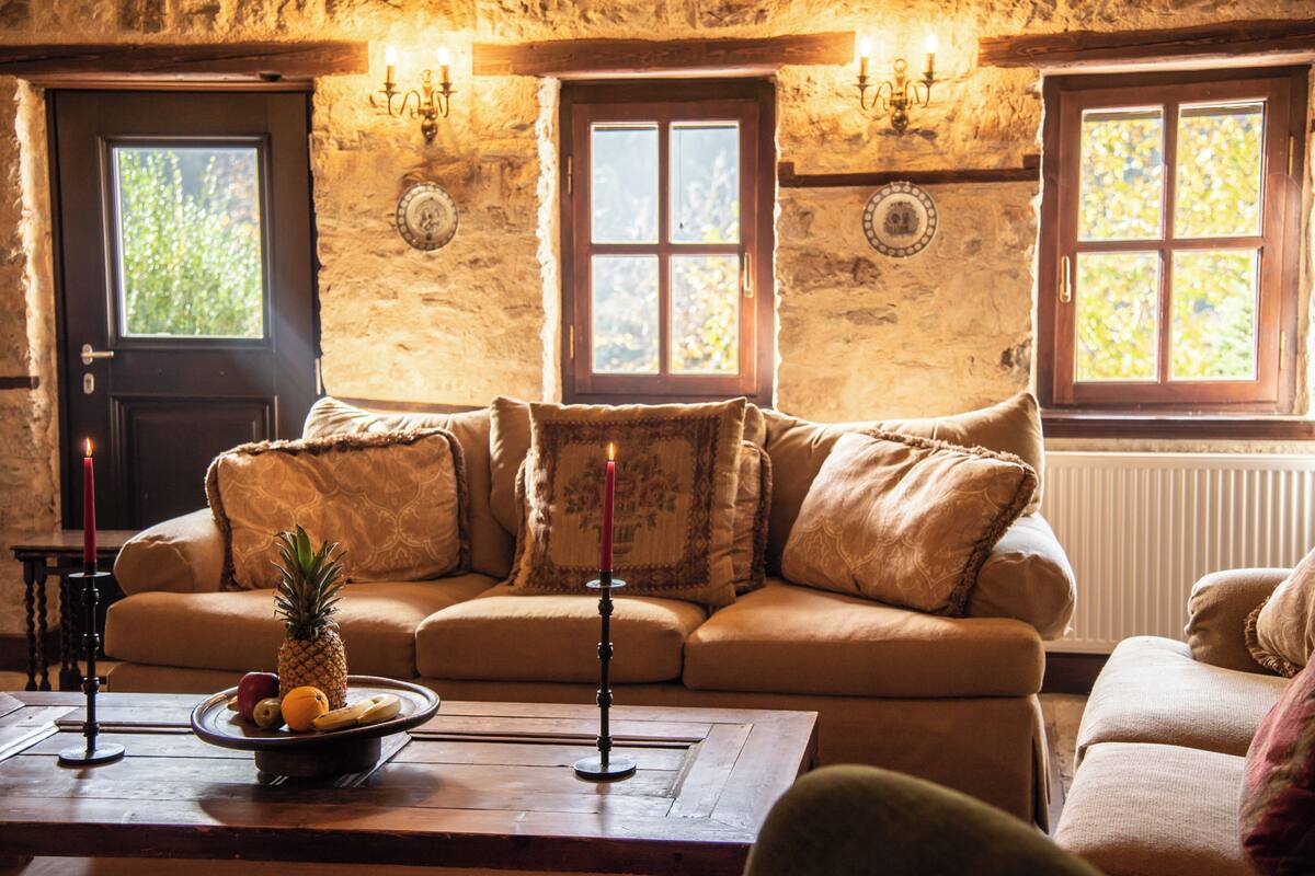 Una habitación acogedora con pared de piedra, puerta de madera y ventana con vistas a árboles. Hay un sofá con cojines y una mesa con frutero y velas.