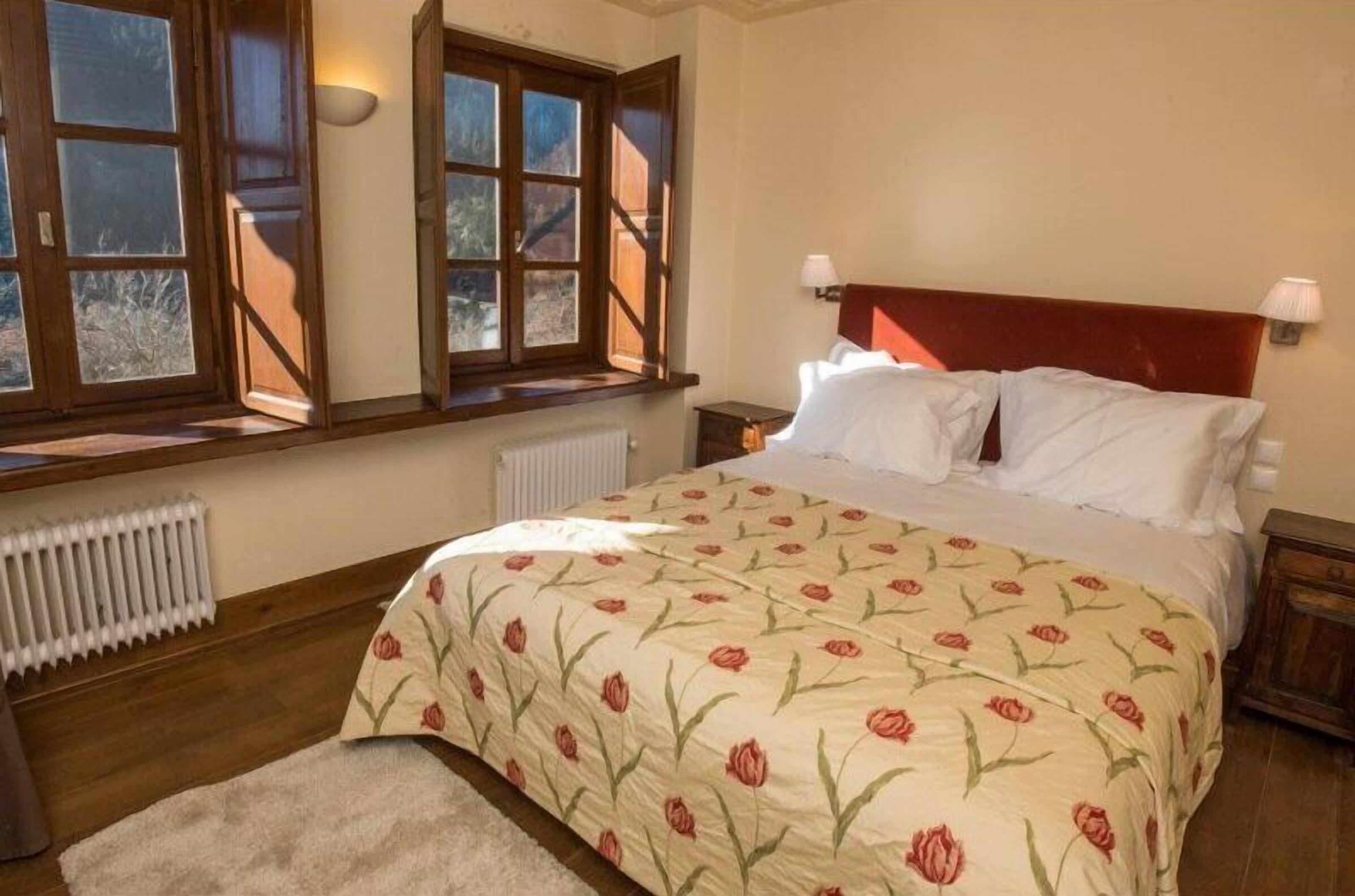 Une chambre avec un grand lit, une tête de lit en bois, deux tables de chevet, un radiateur et une fenêtre donnant sur des arbres.
