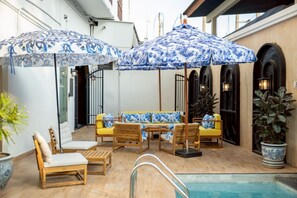 A poolside area with wooden lounge chairs, a yellow sofa, and blue patterned umbrellas. - Metropole, Bangkok, a Tribute Portfolio Hotel (Bangkok)