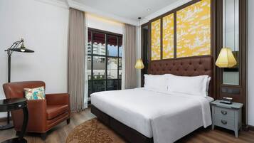 Ein Hotelzimmer mit einem großen Bett, einem braunen Ledersessel, einem kleinen Beistelltisch, einem Nachttisch, einer Lampe und einem Fenster mit Vorhängen.