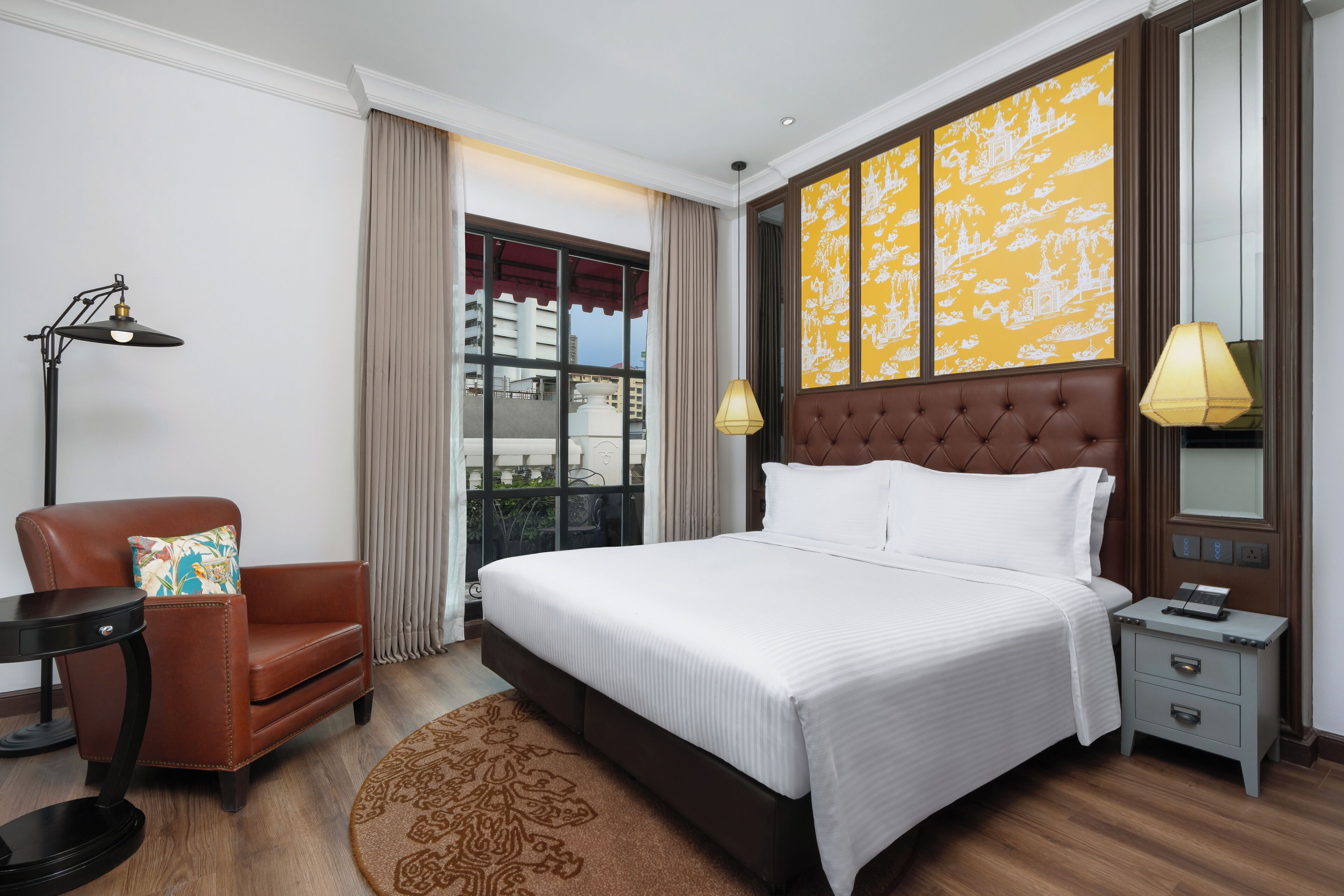 Una habitación de hotel con una cama grande, un sillón de cuero marrón, una mesita auxiliar, una lámpara y una ventana con cortinas.