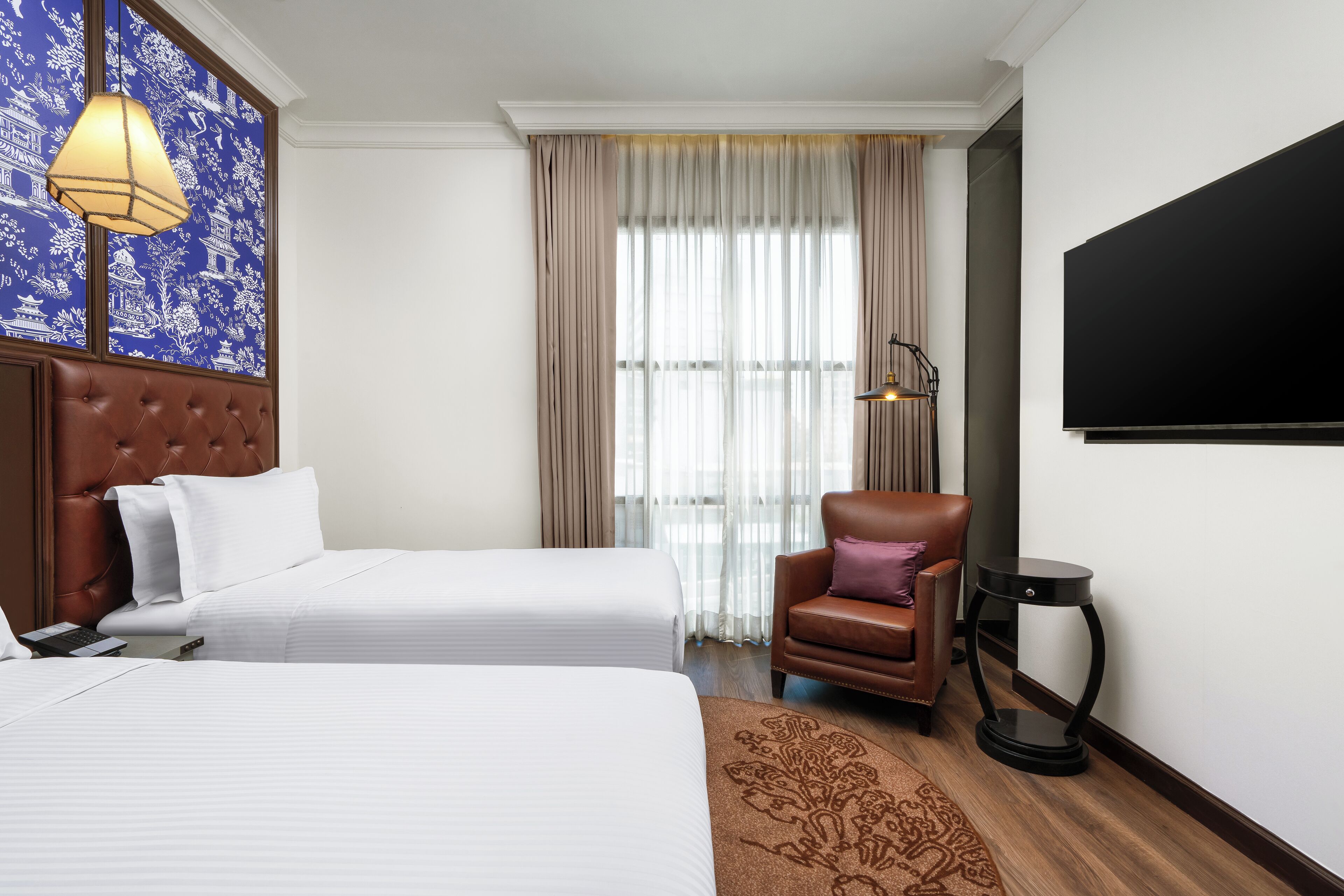 Een hotelkamer met twee bedden, een bruine leren fauteuil, een klein rond tafeltje, een televisie en een kamerkleed met een patroon.