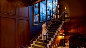 A grand staircase with a woman standing at the top, surrounded by framed pictures on the walls. - Metropole, Bangkok, a Tribute Portfolio Hotel (Bangkok)