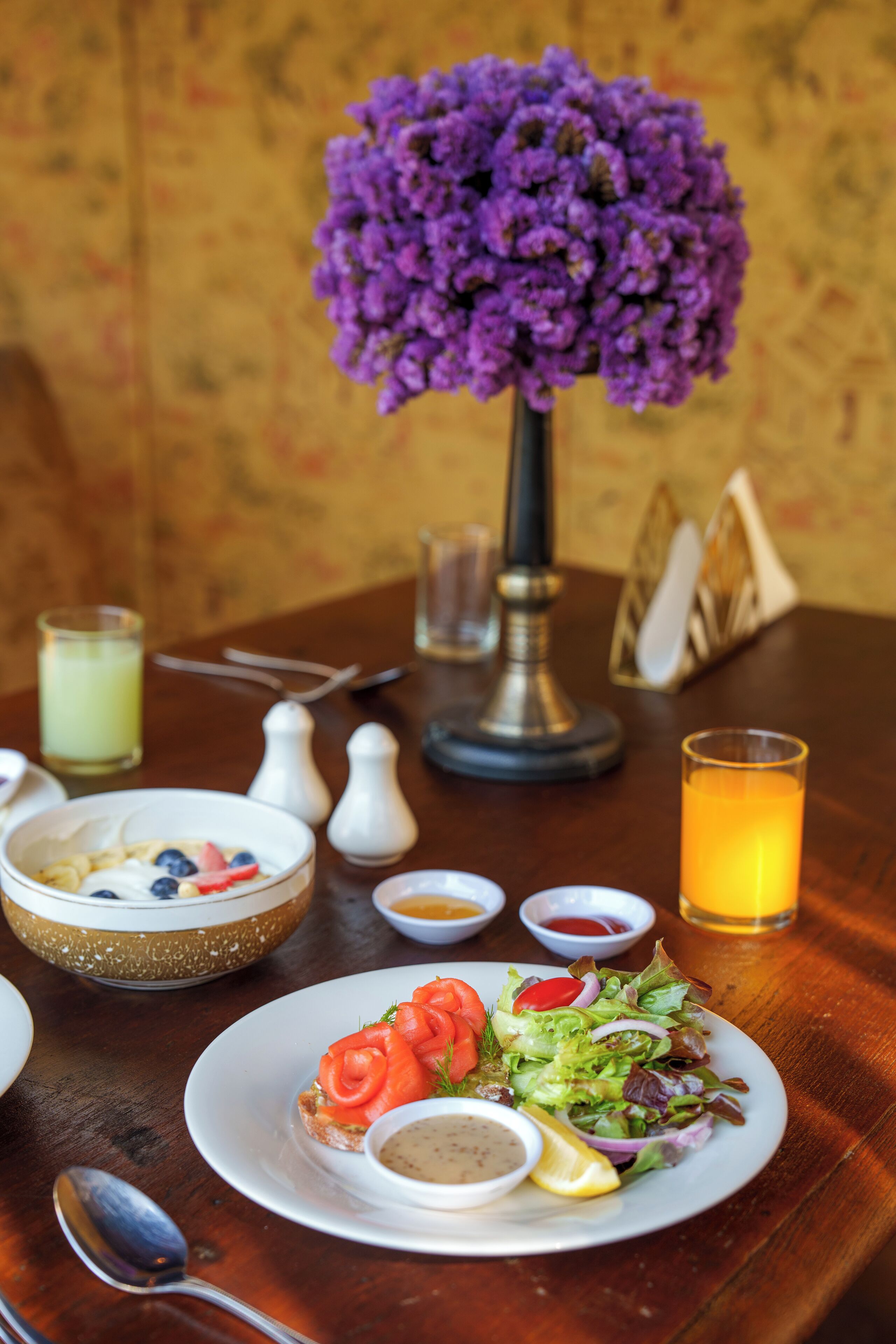 A dining table with a plate of food, a glass of orange juice, and a vase of purple flowers.