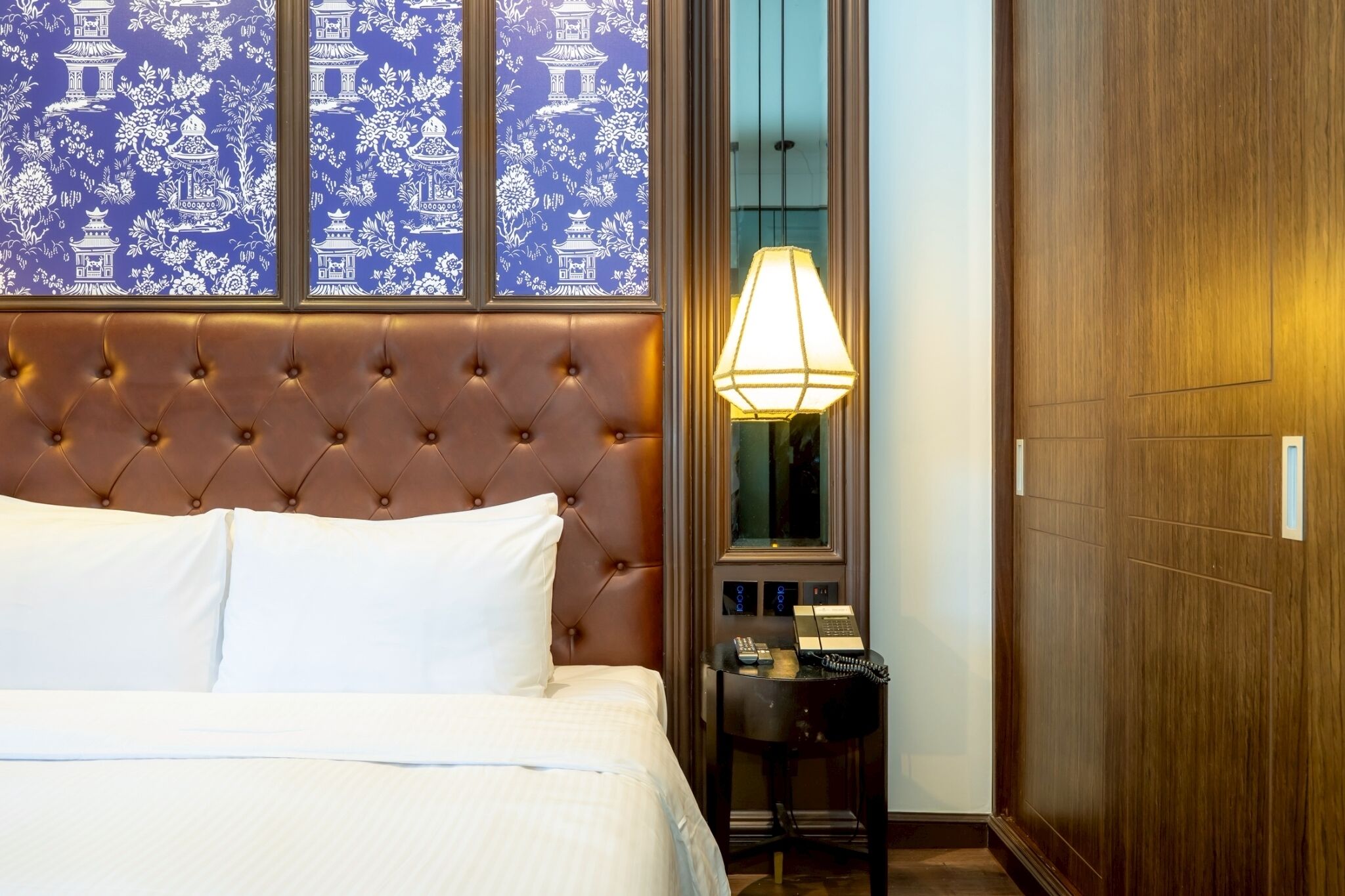 A hotel room with a tufted headboard, a large window with blue floral wallpaper, a bedside lamp, and a wooden wardrobe.