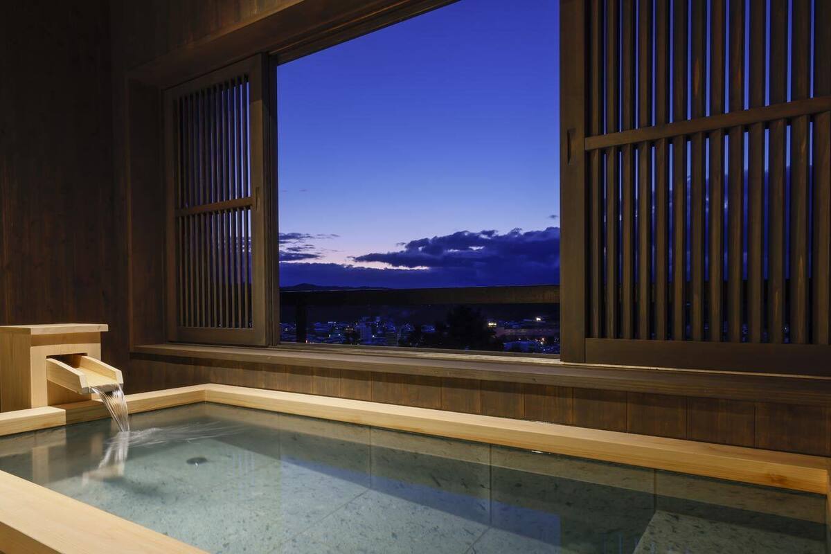Un onsen japonés tradicional con interiores de madera, una bañera de madera y vistas a un paisaje urbano al atardecer.