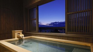A traditional Japanese onsen with a wooden interior, a wooden bathtub, and a view of a cityscape at dusk. - Yoshidaya Sannokaku (Kaga)
