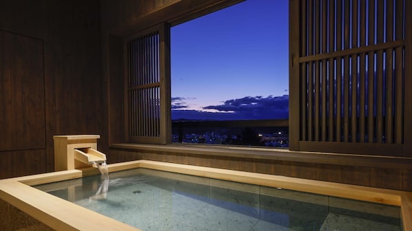 A traditional Japanese onsen with a wooden interior, a wooden bathtub, and a view of a cityscape at dusk.