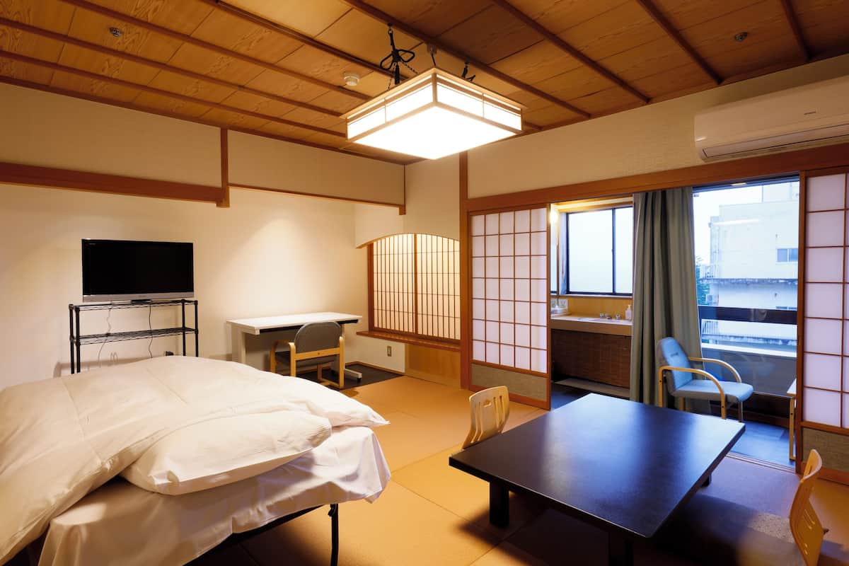 Una habitación tradicional japonesa con suelo de tatami, una cama, un escritorio, un televisor y una puerta corrediza que da a un balcón.