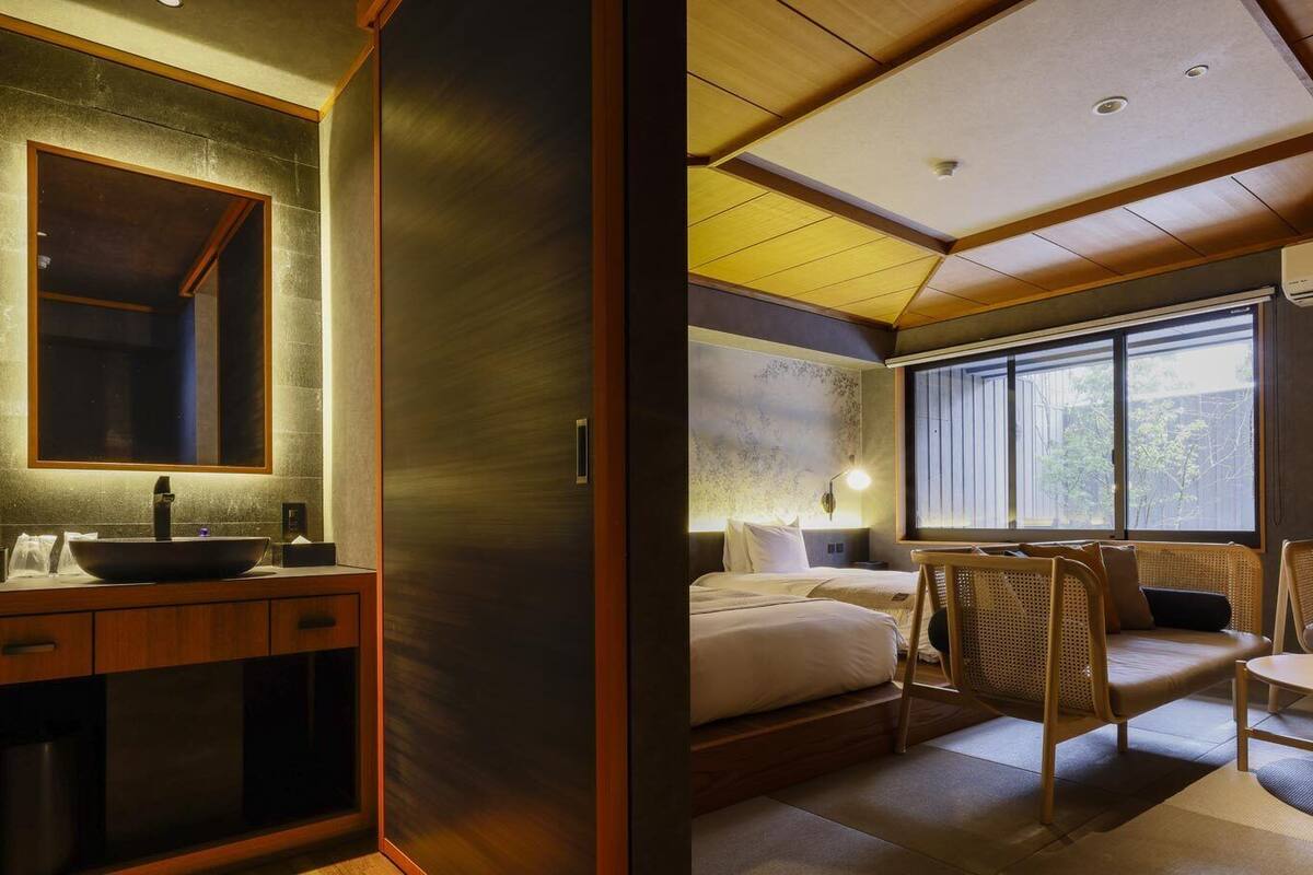 Una habitación de hotel moderna con una cama grande, una zona de estar con una mesa y sillas, un ventanal amplio y un baño con lavamanos y espejo.