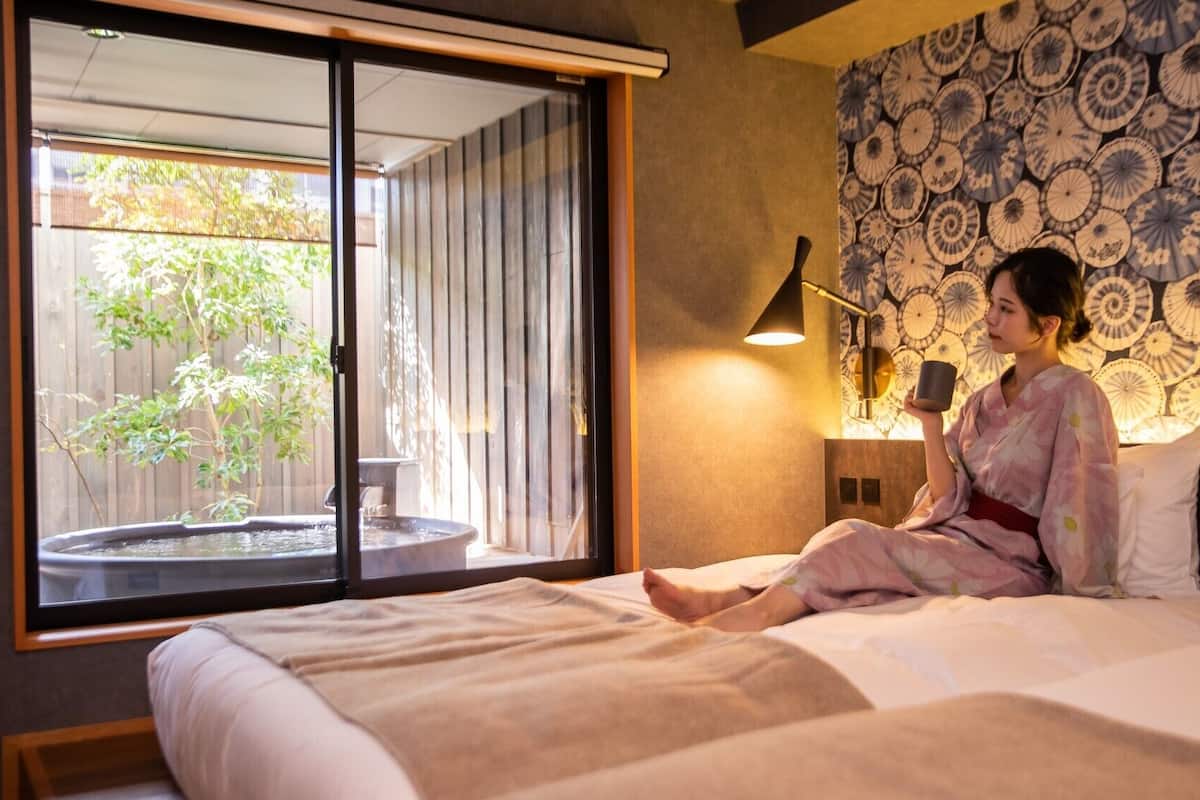 Una habitación con cama, ventana con vista a follaje, pared con diseño decorativo y mesita de noche con lámpara.