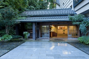 Un edificio japonés tradicional con tejado de tejas, entrada empedrada y un pequeño jardín con plantas y una fuente de agua.