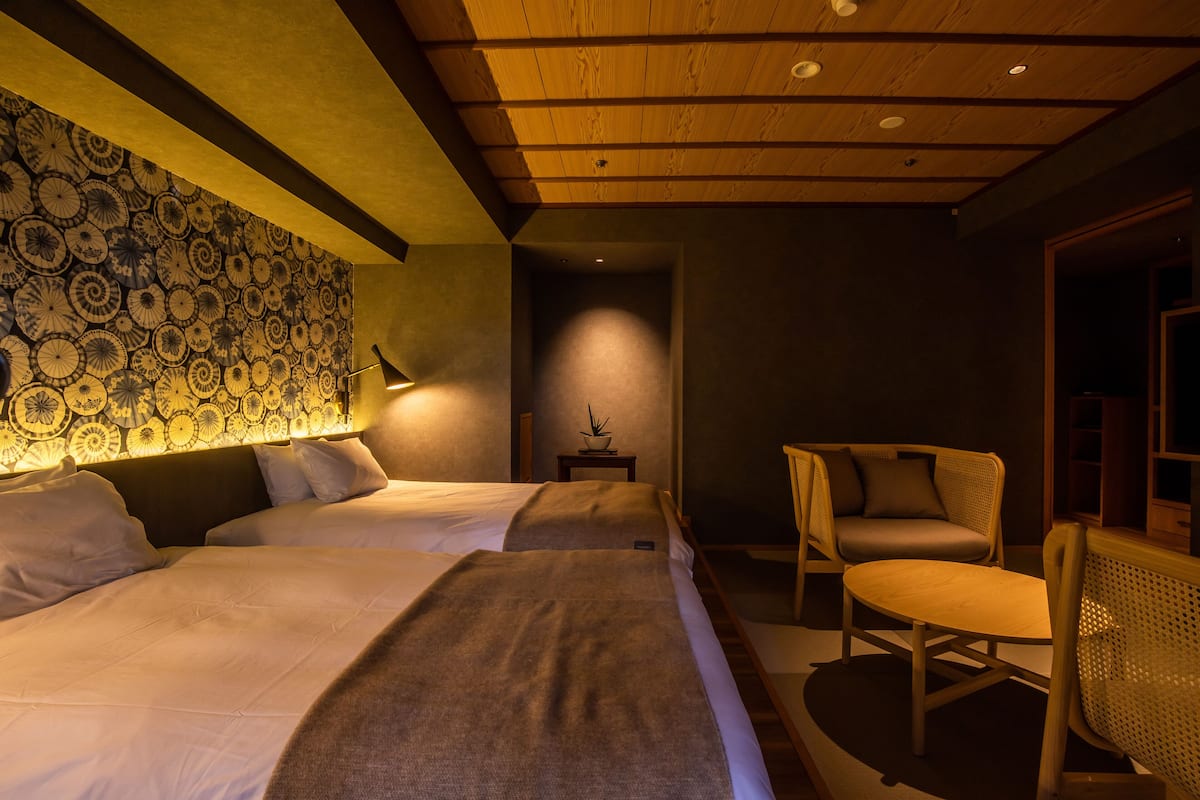 Habitación de hotel con dos camas, una mesita, una silla y un papel tapiz estampado.