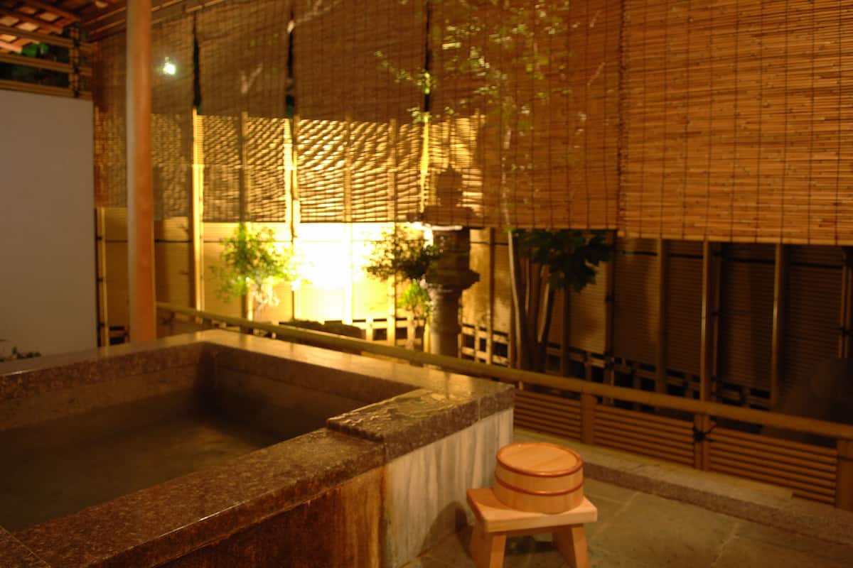 Un onsen japonés tradicional con una fuente termal de piedra, asientos de madera y biombos de bambú.