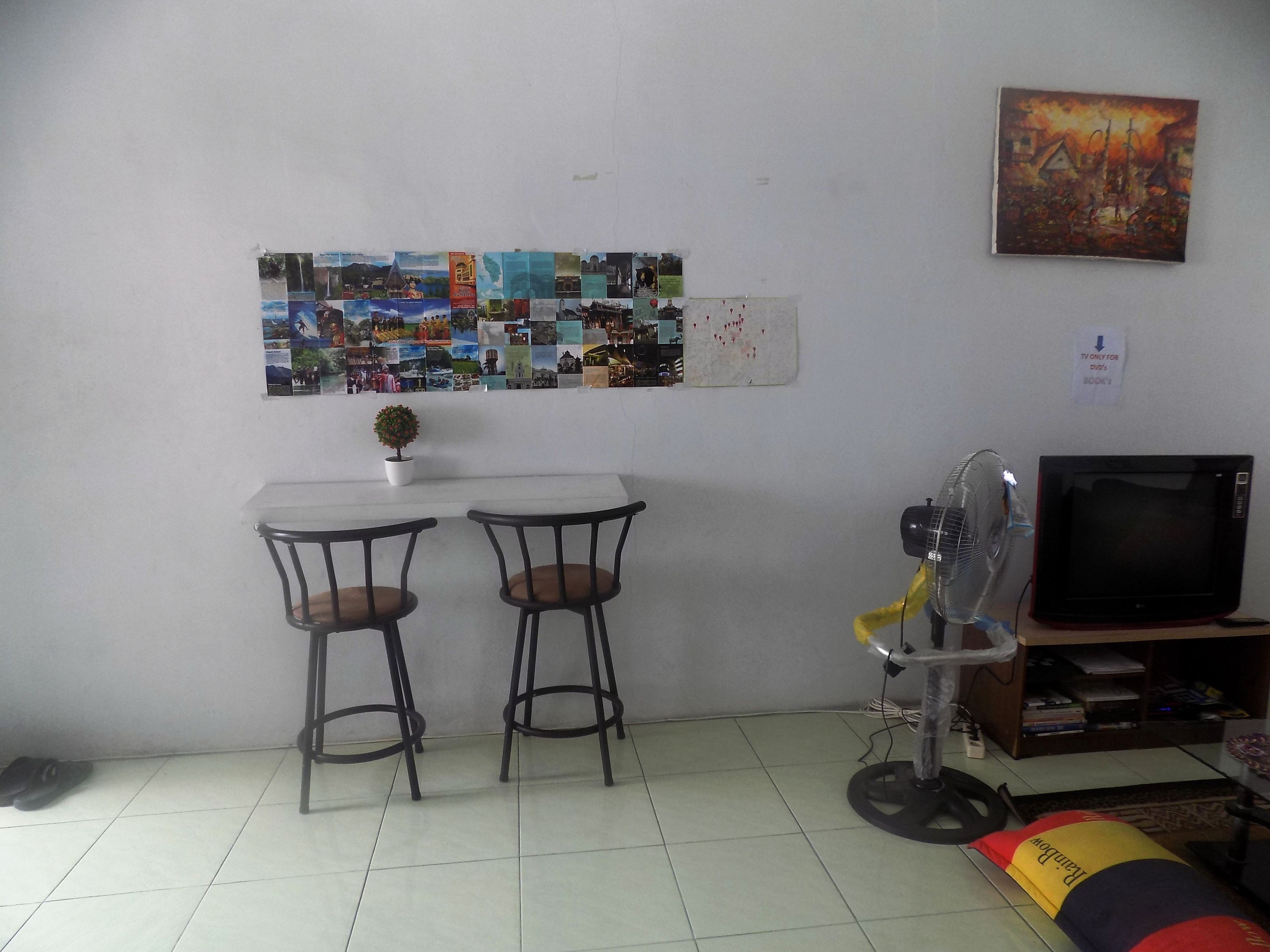 A small room with two tables and chairs, a wall-mounted TV, a fan, and a bulletin board with various pictures.