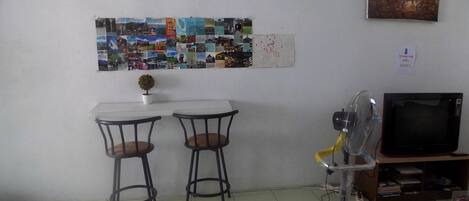 A small room with two tables and chairs, a wall-mounted TV, a fan, and a bulletin board with various pictures.
