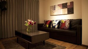 A modern living room with a dark brown coffee table, a vase with flowers, a black leather sofa with colorful cushions, and a wall art piece above the sofa. - Chelsea24 Kyoto (Kyoto)