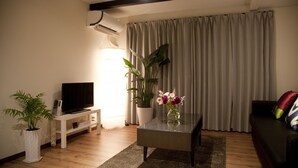 A living room with a flat-screen TV on a white stand, a glass coffee table with a vase of flowers, a potted plant, and a sofa with a colorful pillow. - Chelsea24 Kyoto (Kyoto)