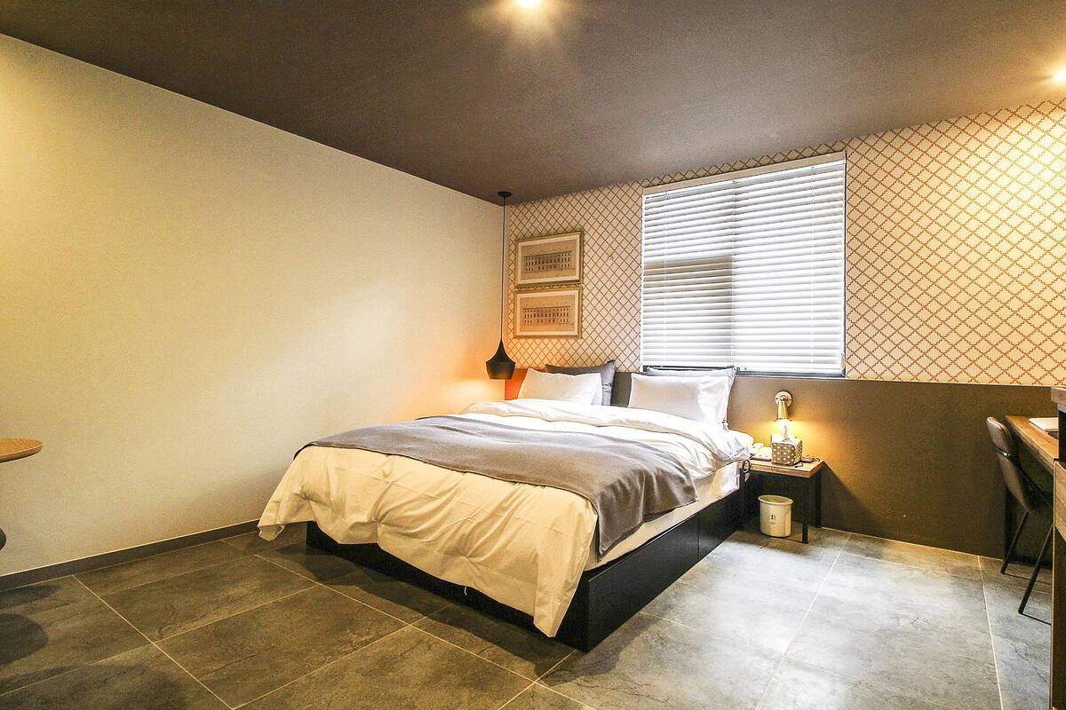 A bedroom with a bed, a nightstand, a lamp, a window with blinds, and a patterned wall.