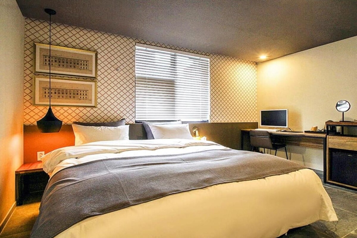 Hotellihuone, jossa on sänky, työpöytä, tuoli, tietokone ja ikkuna, jossa on sälekaihtimet.