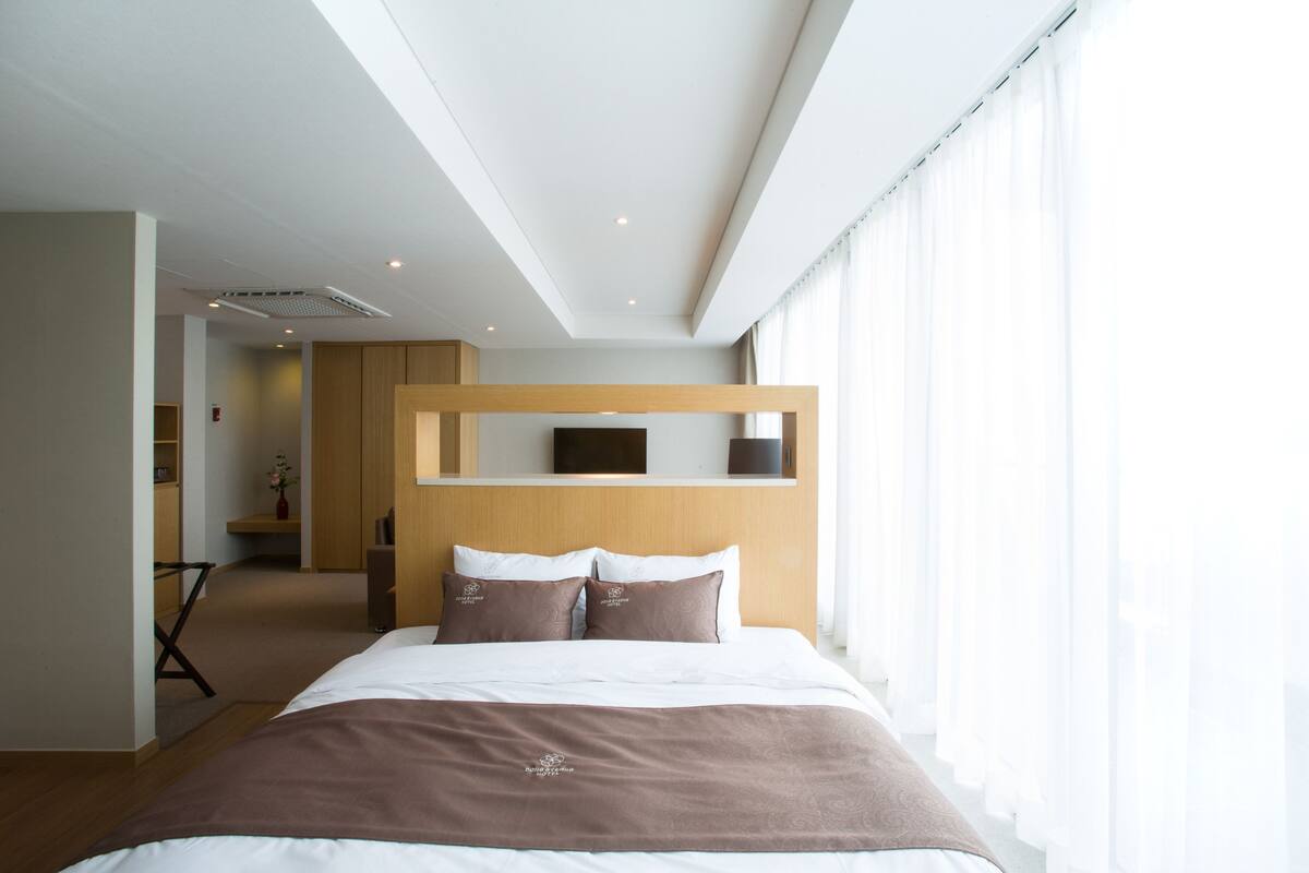 Una habitación de hotel moderna con una cama grande, cabecera de madera, televisor de pantalla plana y vistas a la ciudad a través de amplias ventanas.