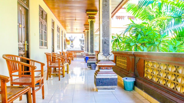 A corridor with wooden chairs and benches, a tiled floor, and a column with a decorative pattern. - Lolo Beach Inn (Legian)