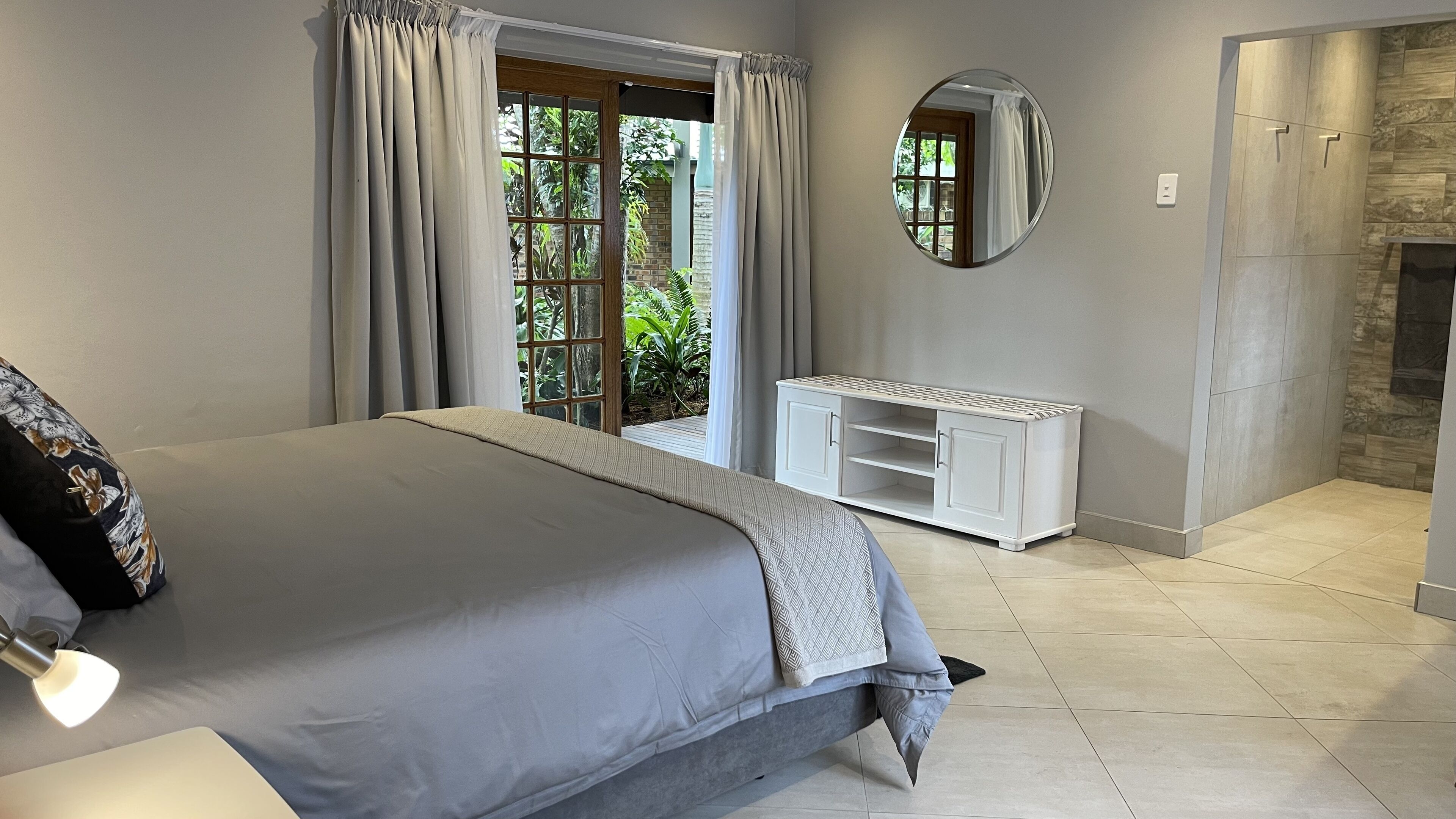 A bedroom with a large bed, a white console table, a mirror, and a view of greenery outside.