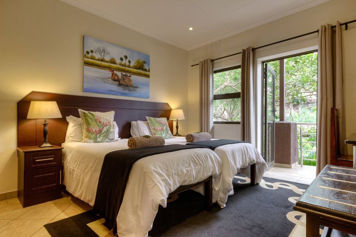 Quarto de hotel com duas camas, uma mesa de cabeceira, um quadro de uma paisagem fluvial e uma janela grande com cortinas.