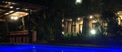 A nighttime swimming pool with clear water reflecting lights, surrounded by lush greenery and a building with a covered patio area.